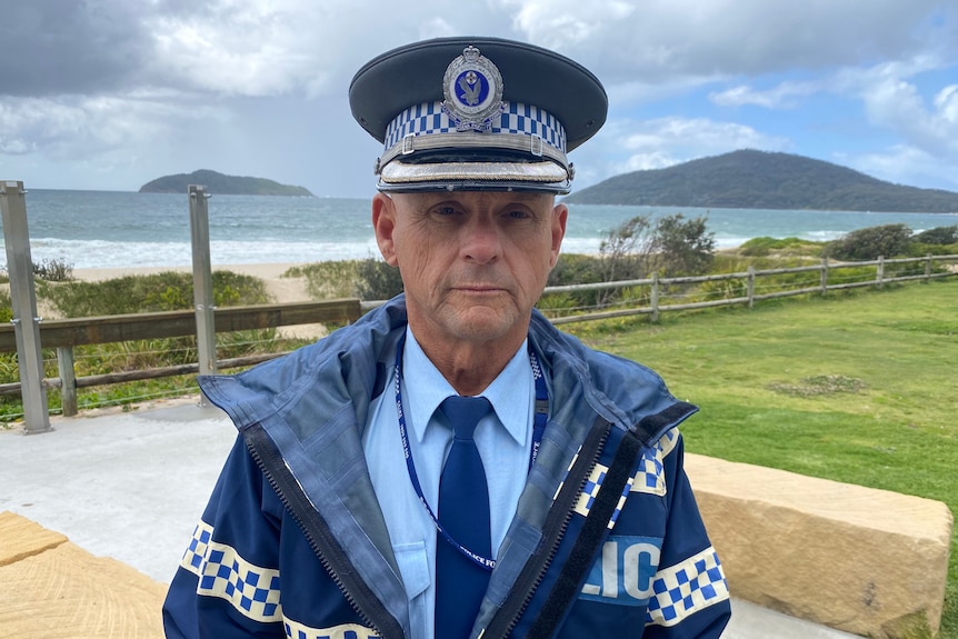 Policeman in uniform looks at camera with serious expression, with beach behind him.