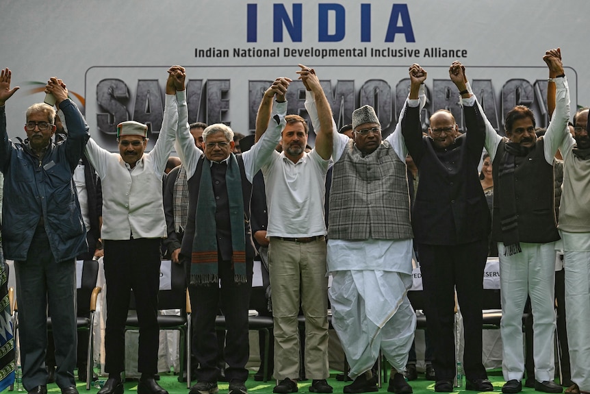 A group of male political leaders raise held hands as they stand in a line in front of a sign that says INDIA.