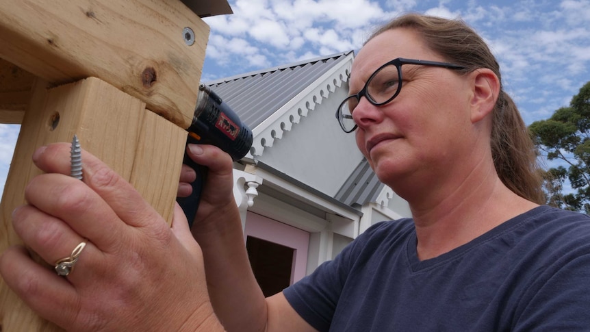 A woman wearing glasses and holding a screw, drilling into some pine timber, with a decorative cubby house in the background.