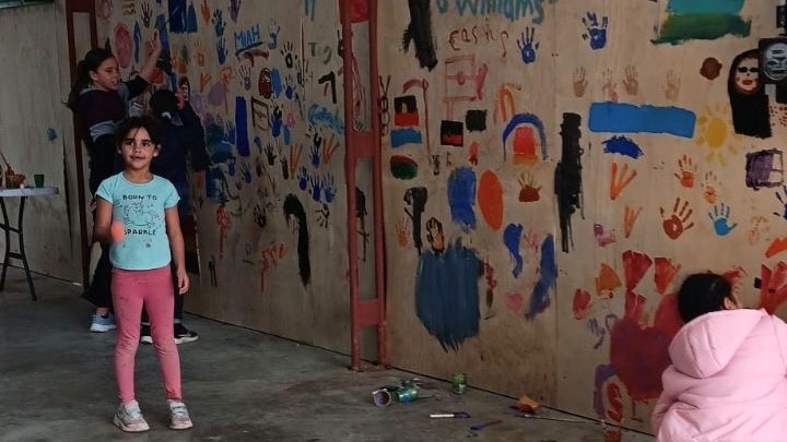 A young Aboriginal girl in pink pants and blue top stands next to a wooden wall with multicoloured paintings