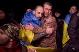 A man draped in a Ukranian flag hugs a man in army fatigues among other released prisoners of war