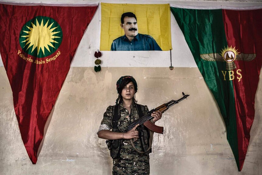 A woman in military fatigues, holding an assault rifle, poses in front of a picture of a man.