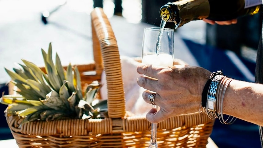 picnic basket in the background,  champagne being poured out of a bottle into a glass in the foreground.