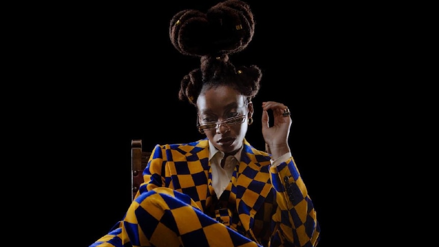 British rapper Little Simz sits in a blue and yellow checkered suit, wears glasses and hair in updo