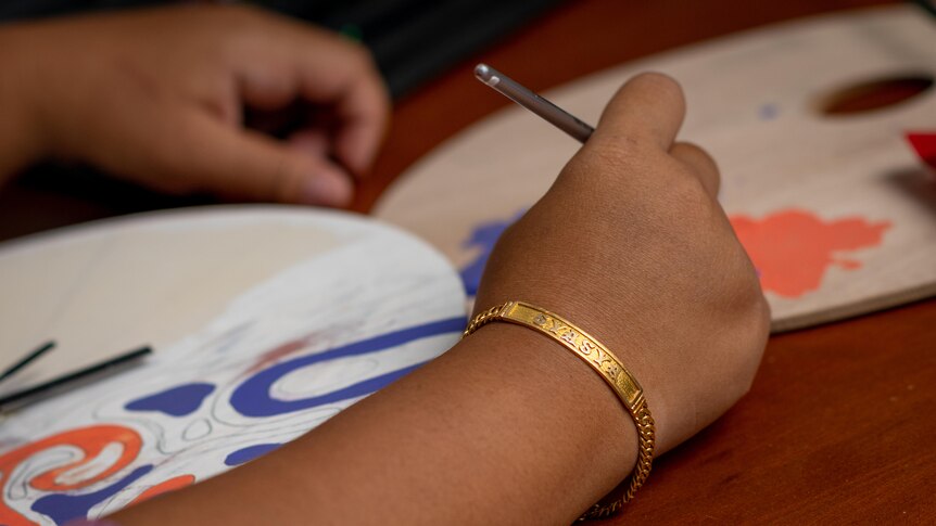 A woman's hand holds a paintbrush over a semi coloured in drawing, on her arm is a gold bracelet that reads Syasya