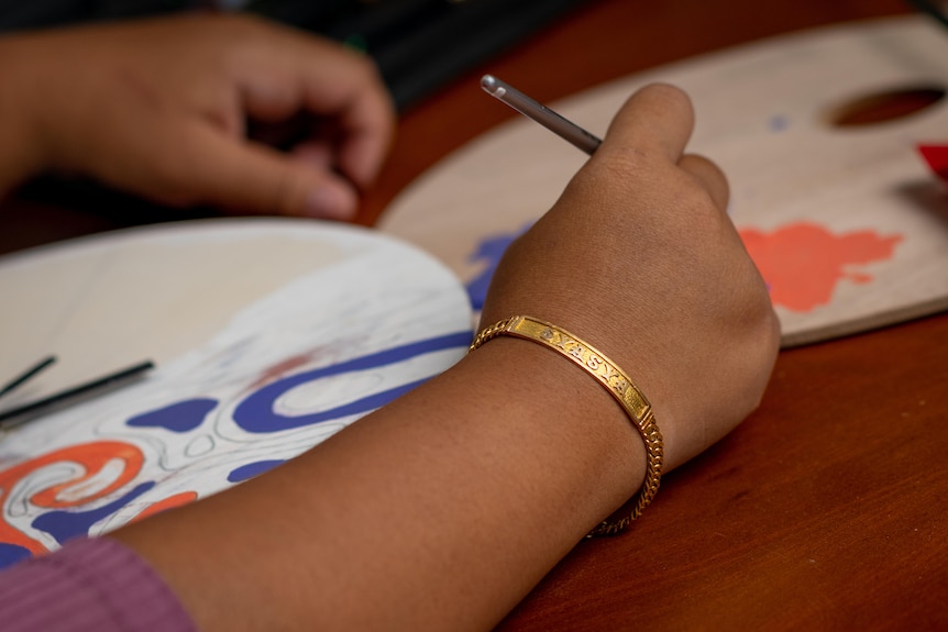 A woman's hand holds a paintbrush over a semi coloured in drawing, on her arm is a gold bracelet that reads Syasya