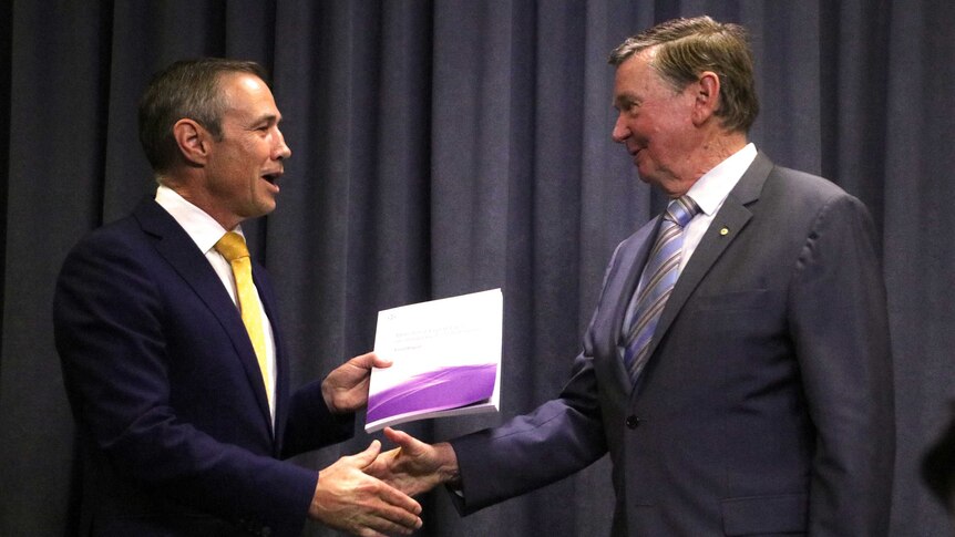 A mid shot of Roger Cook and Malcolm McCusker standing inside shaking hands as Mr Cook holds a report.