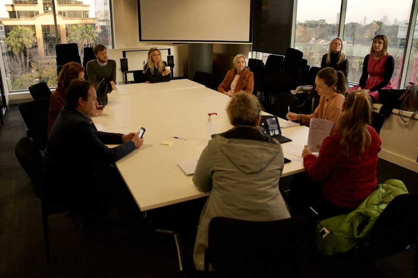 Ten people, mostly women, sit around a board table in a room with big glass windows and views of the city.