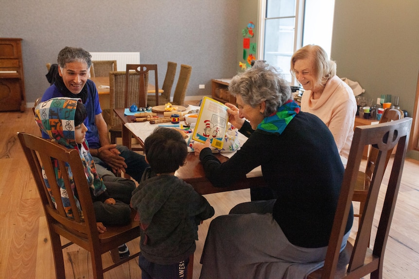 Two young kids are read a story by an older woman, while other adults watch on.