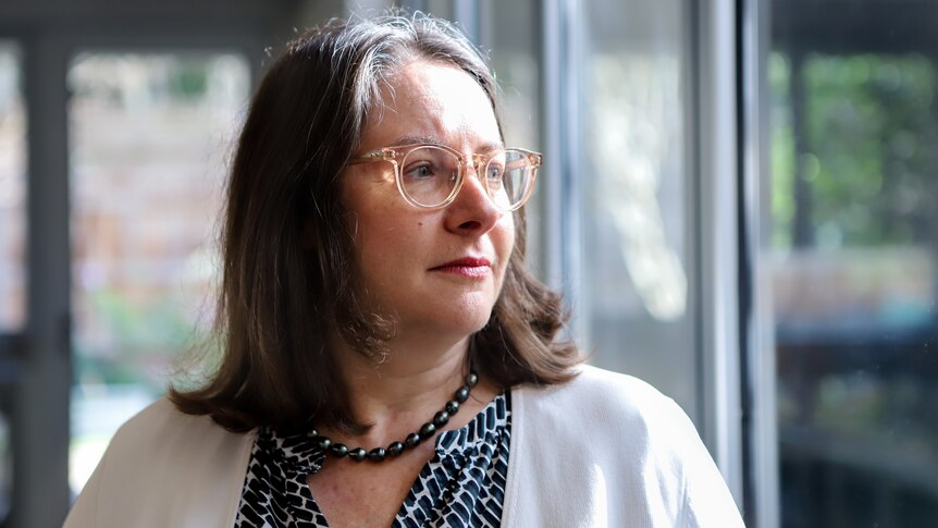 Woman wearing glasses with brown hair and white cardigan stares out glass window from inside home kitchen