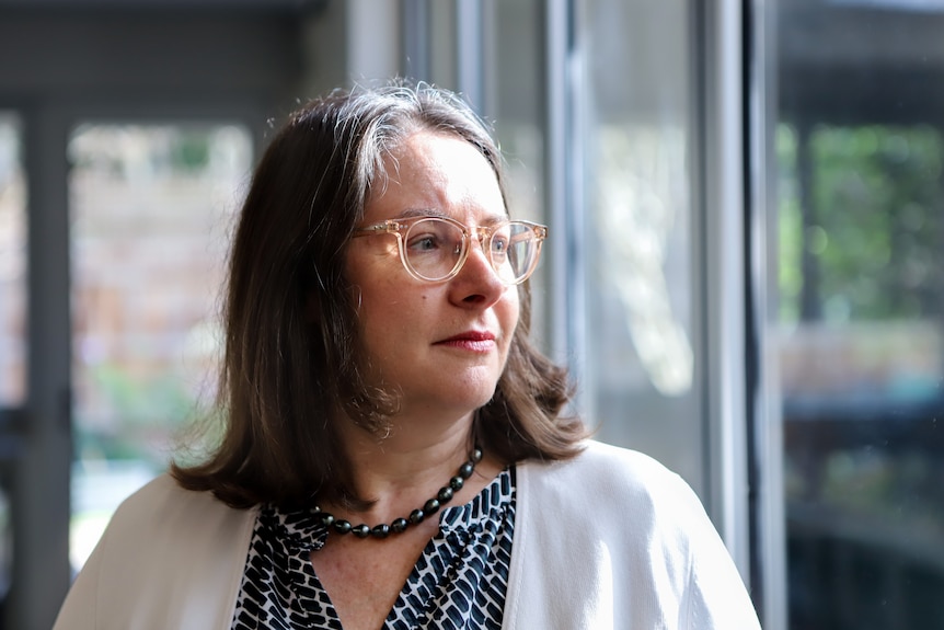Woman wearing glasses with brown hair and white cardigan stares out glass window from inside home kitchen