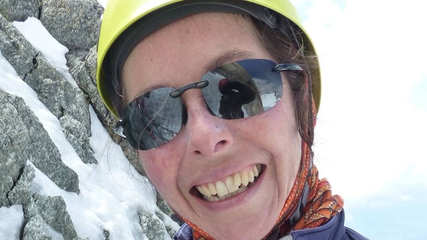 A smiling woman stands on a snowy mountain wearing a yellow helmet and purple jacket.