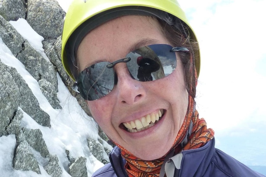 A smiling woman stands on a snowy mountain wearing a yellow helmet and purple jacket.