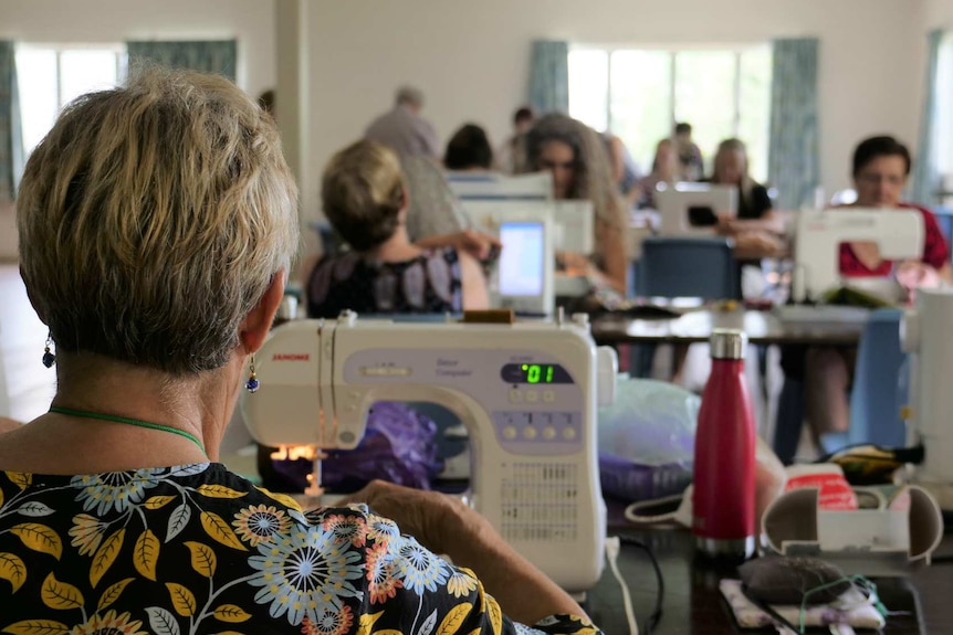 The back of a woman's head at a sewing machine with more people behind sewing machines in the background