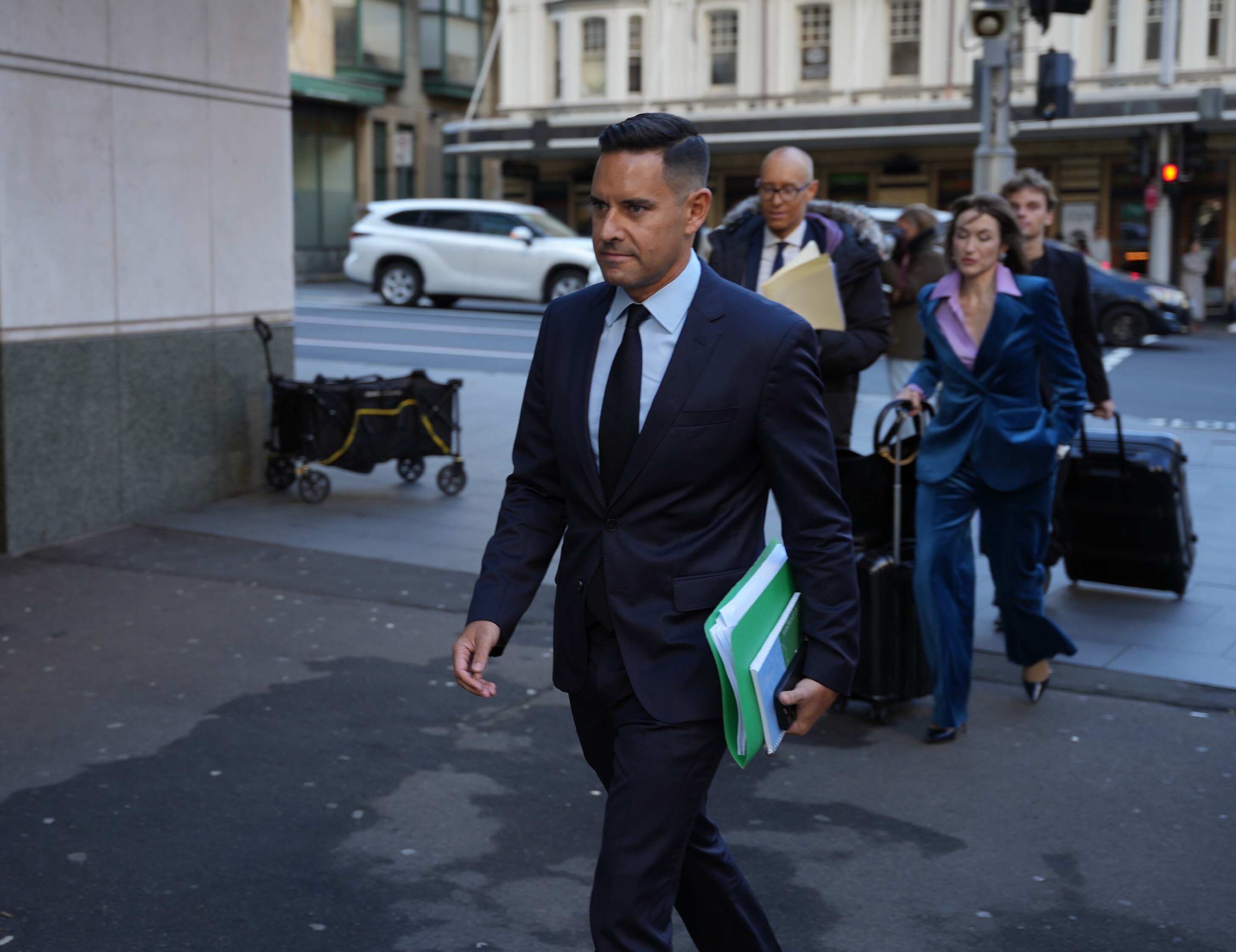Alex Greenwich holds binder with people walking behind him