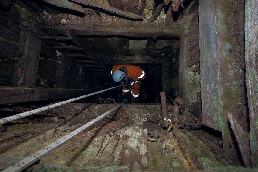 a man abseils into a mine shaft, which has walls lined with old wood.