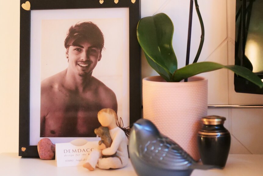 A bird shaped urn sits next to a framed photo of a young shirtless man with a moustache and goatee