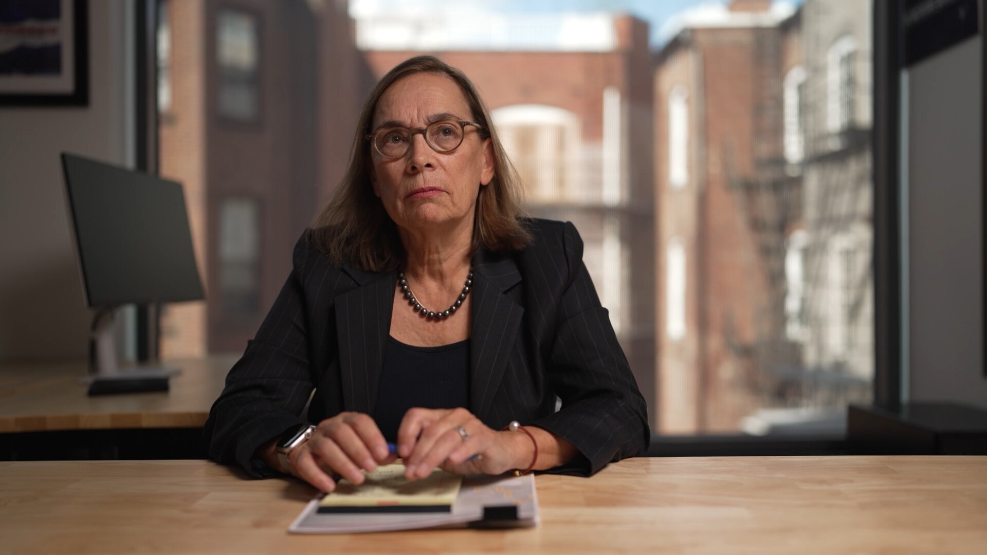 A woman wearing glasses and black blazer sits at a wooden desk in an office. A brick building is seen through the window.