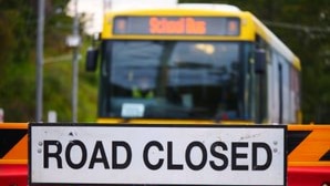 A 'road closed' sign attached to red barricades, a yellow school bus approaches the closure