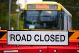 a 'road closed' sign attached to red barricades, a yellow school bus approaches the closure
