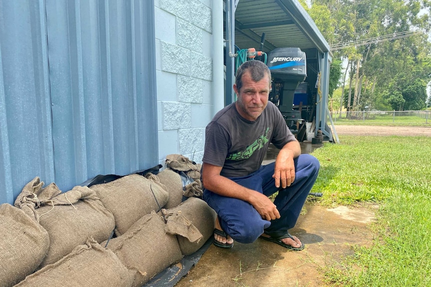 Man kneeling on cement, in front of home, which is lined with sandbags.