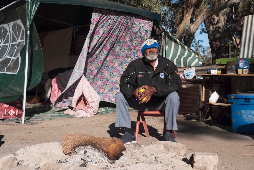An older Aboriginal man sits by an open fire in front of a tent and outdoor kitchen during the day.
