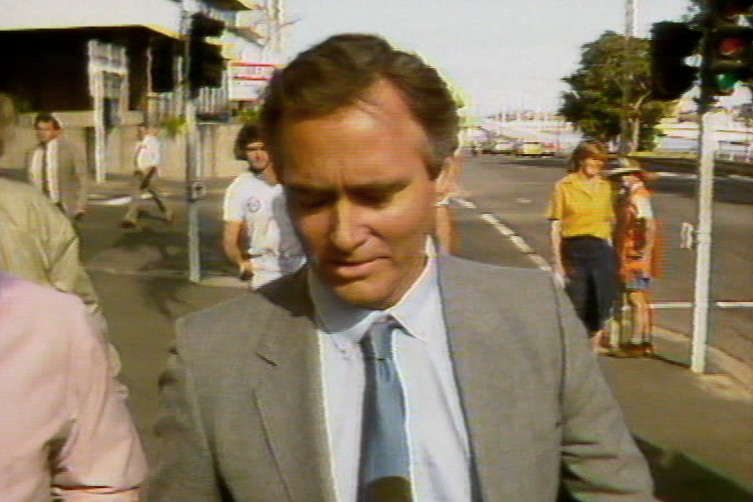 A man with brown hair wearing a grey suit walking along the sidewalk during the day.