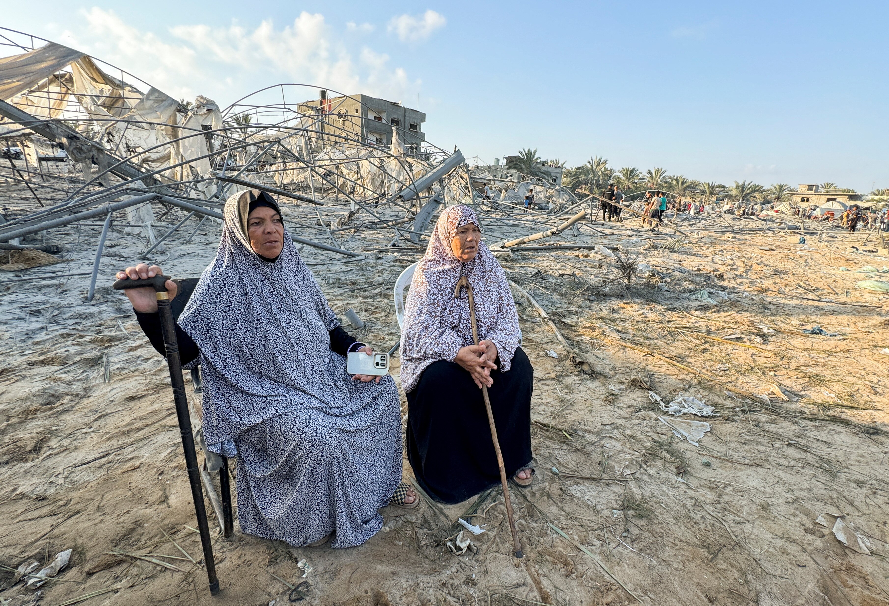Two Palestinian women sit with walking sticks on a chair in the middle of debris