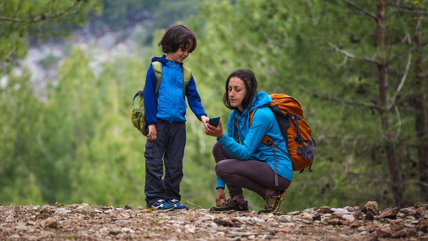 A mother crouches to show her young son how to use her smartphone’s GPS while they are on a bush trail.