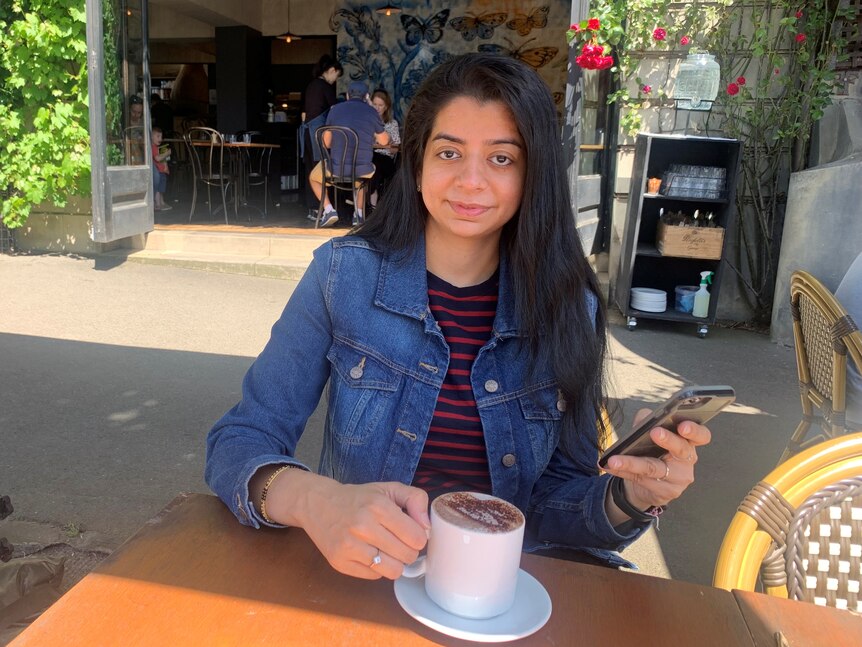 A young Indian woman, straight hair lose, wears denim jacket, striped t-shirts, looks smilingly at phone in hand, drinks coffee.