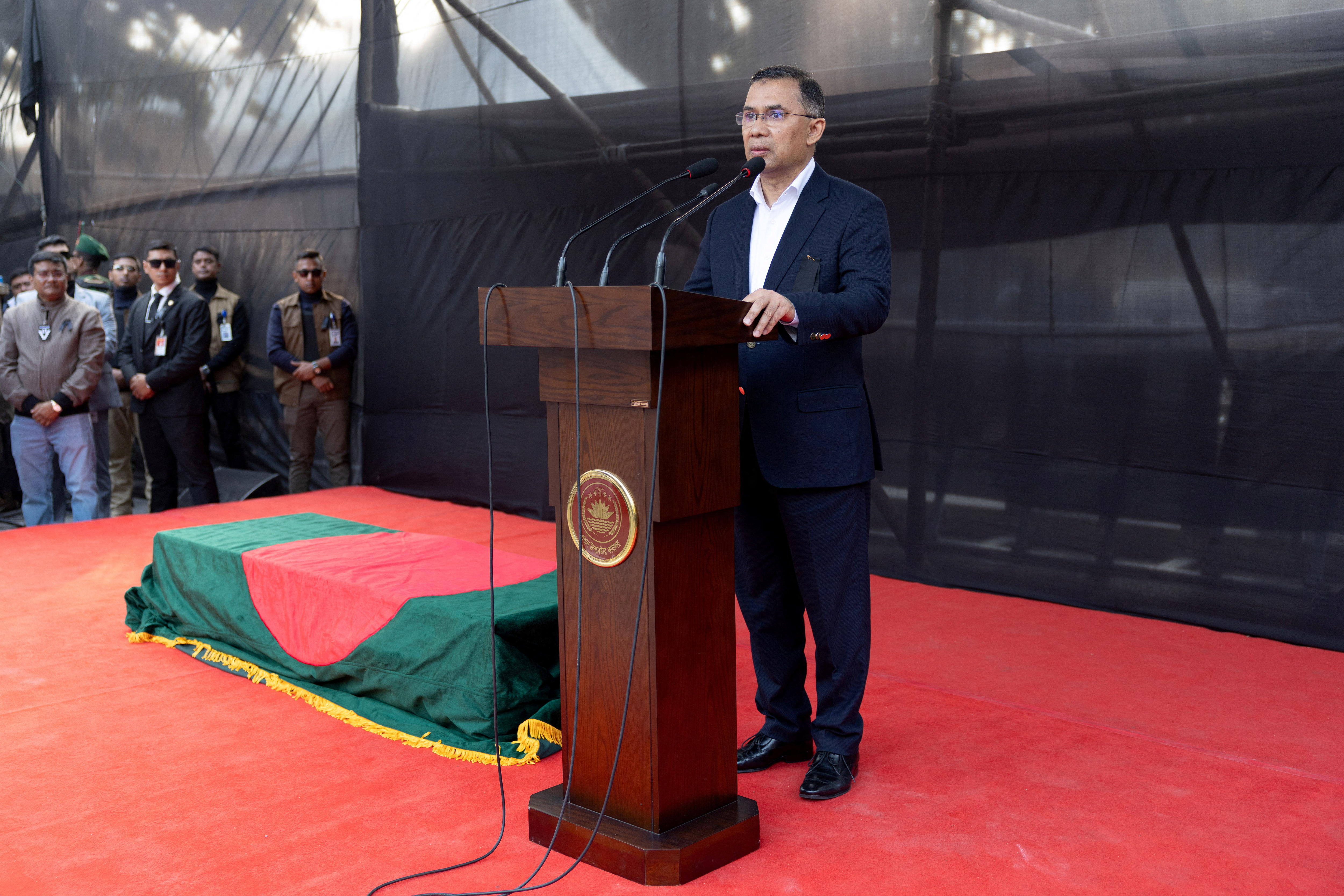 Un hombre con traje azul marino y camisa de cuello abierto da un discurso en el escenario junto a un ataúd cubierto con una bandera verde con un círculo rojo.