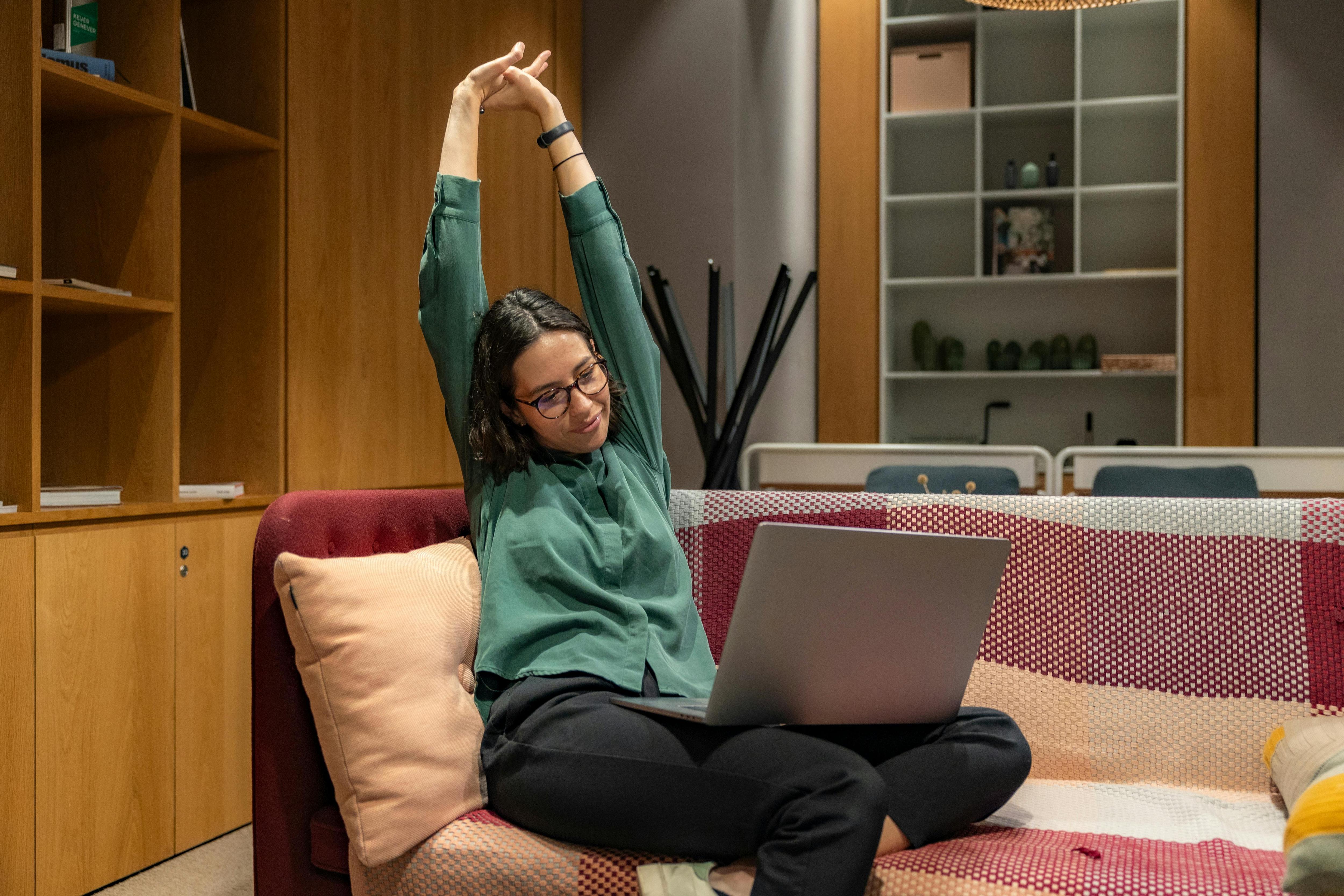 A woman sits on a couch with a laptop stretching
