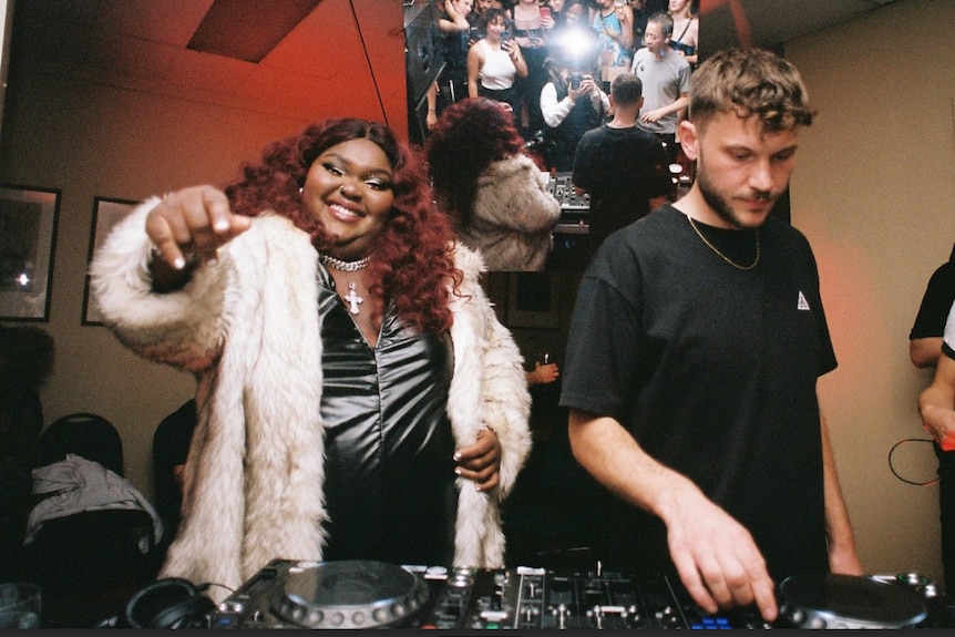 A man in a black shirt and a women in a white coat stand in front of DJ decks. 