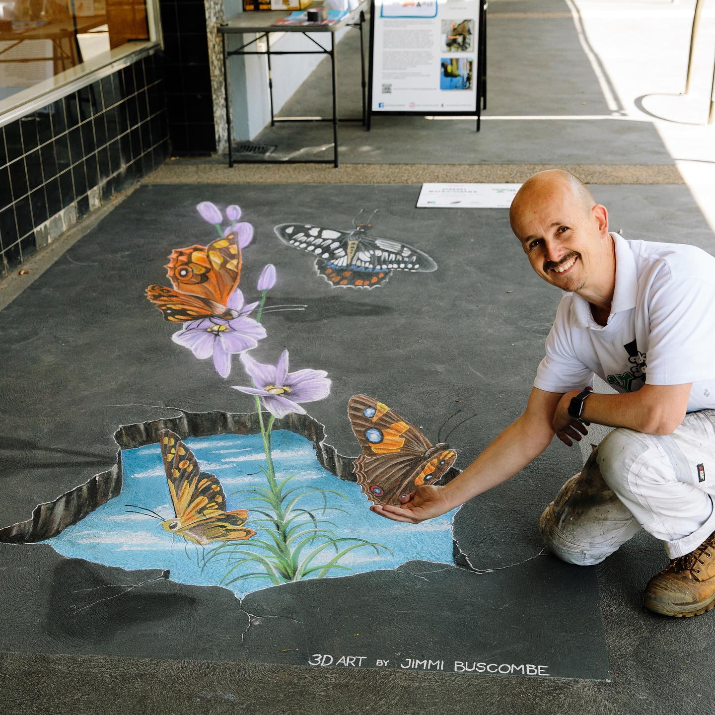 A 3D chalk drawing of a pond with flowers and butterflies on footpath, a man kneels so a butterfly looks perched on his hand.