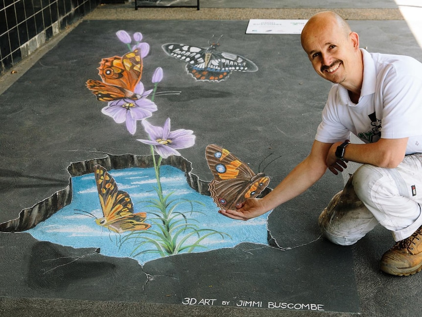 A 3D chalk drawing of a pond with flowers and butterflies on footpath, a man kneels so a butterfly looks perched on his hand.