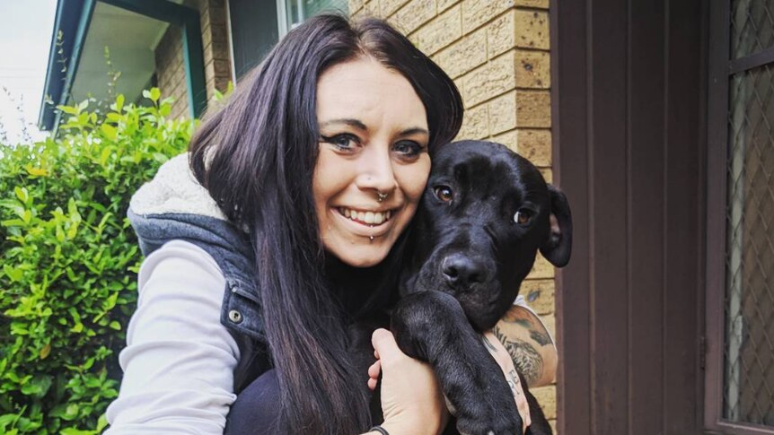 A young woman smiles at the camera while cuddling a big black dog.