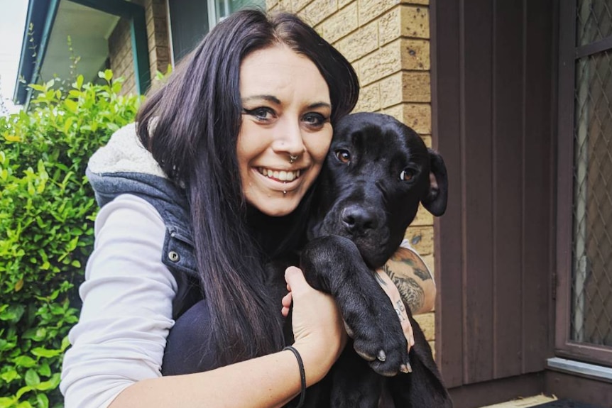 A young woman smiles at the camera as she hugs a large black dog.