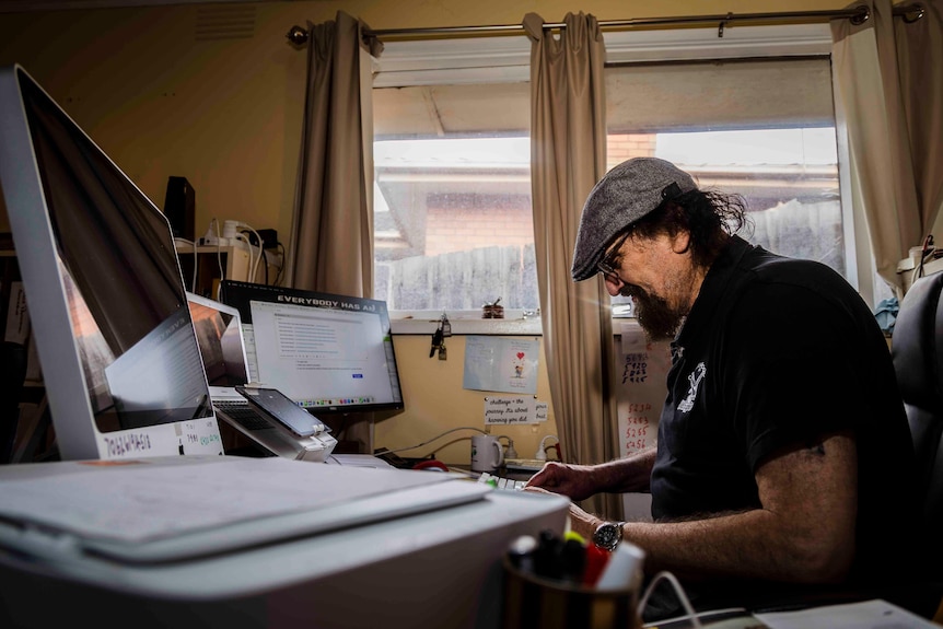 A man wearing a cap and a black shirt at a desk looking at a screen.