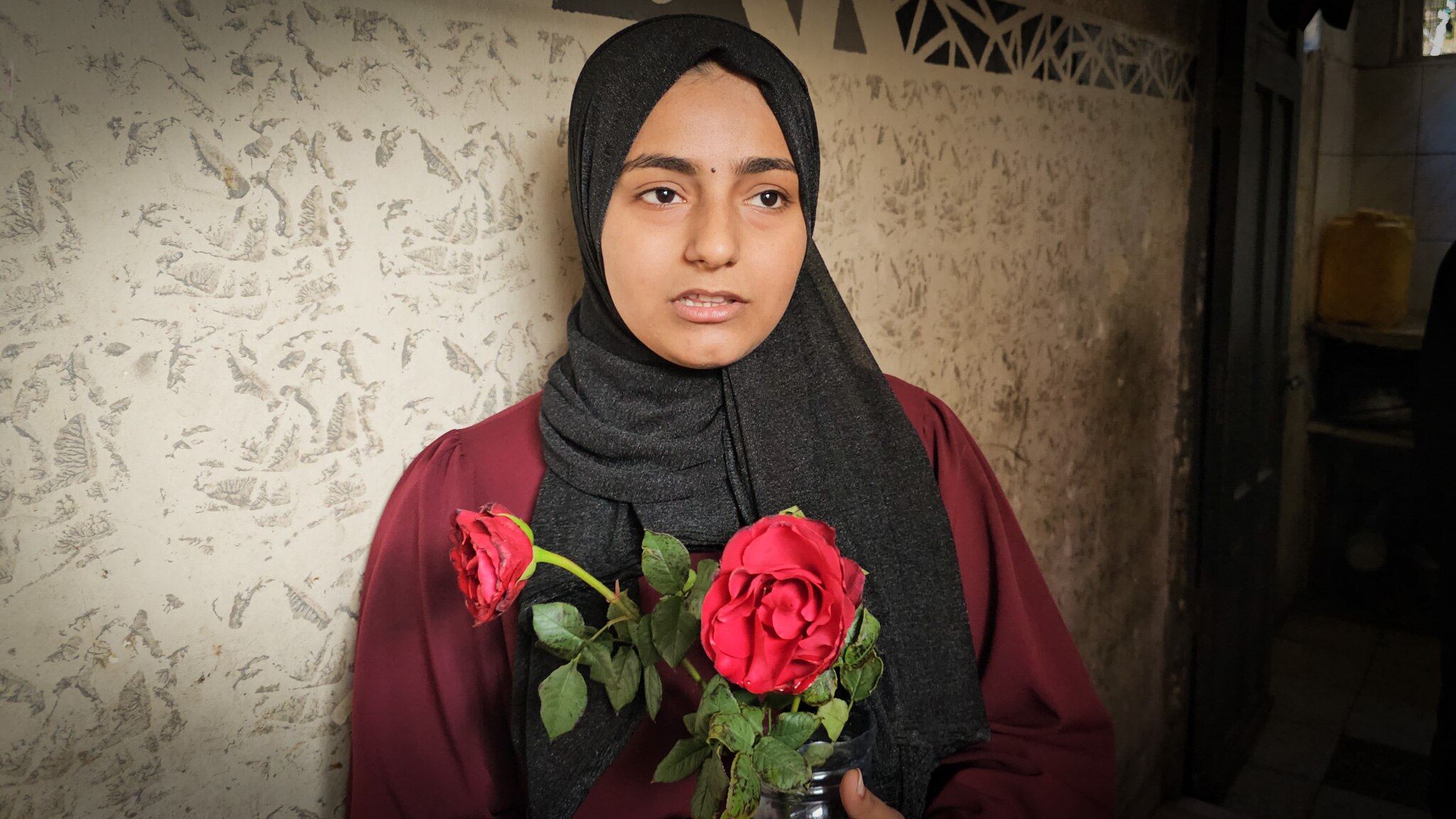 A close up of a girl wearing a black hijab and clutching two red roses to her chest.