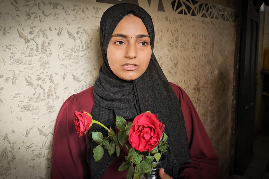 A close up of a girl wearing a black hijab and clutching two red roses to her chest.