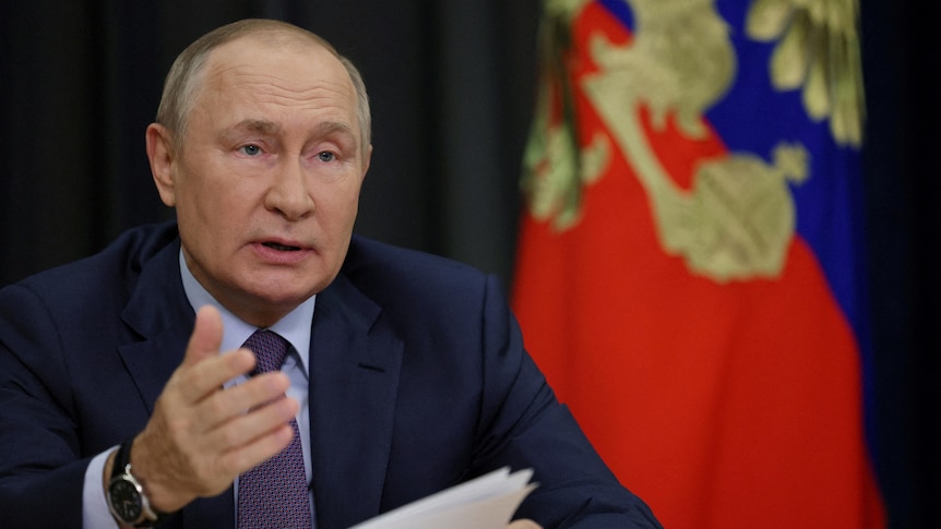 A man in suit raises his right hand as he reads from papers before a Russian flag.