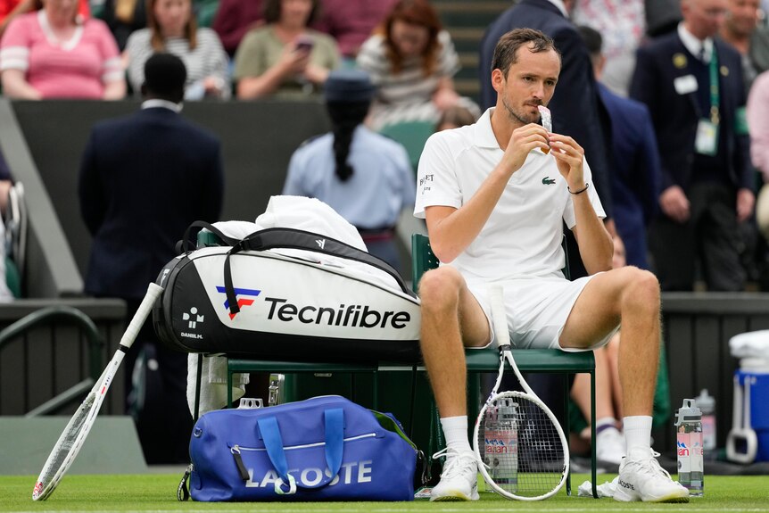 A tennis player sits on his chair at courtside at Wimbledon, eating an energy gel from a tube.