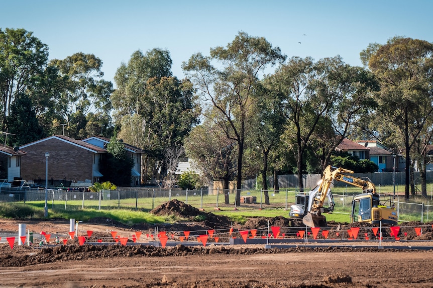 A construction site in the foreground, with older public housing visible through the trees behind.