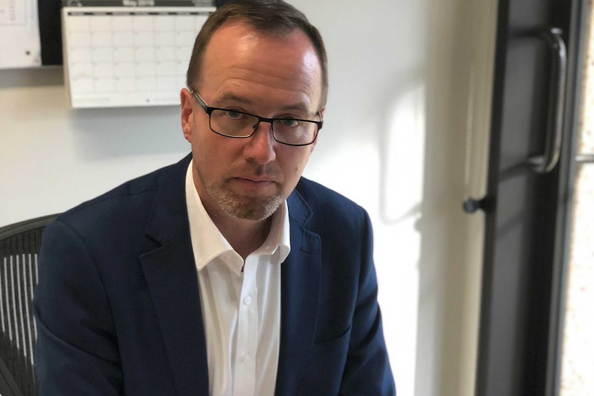 Greens MP Dave Shoebridge sits at a desk in an office wearing a blue suit, holding a pen and with a computer in front of him.