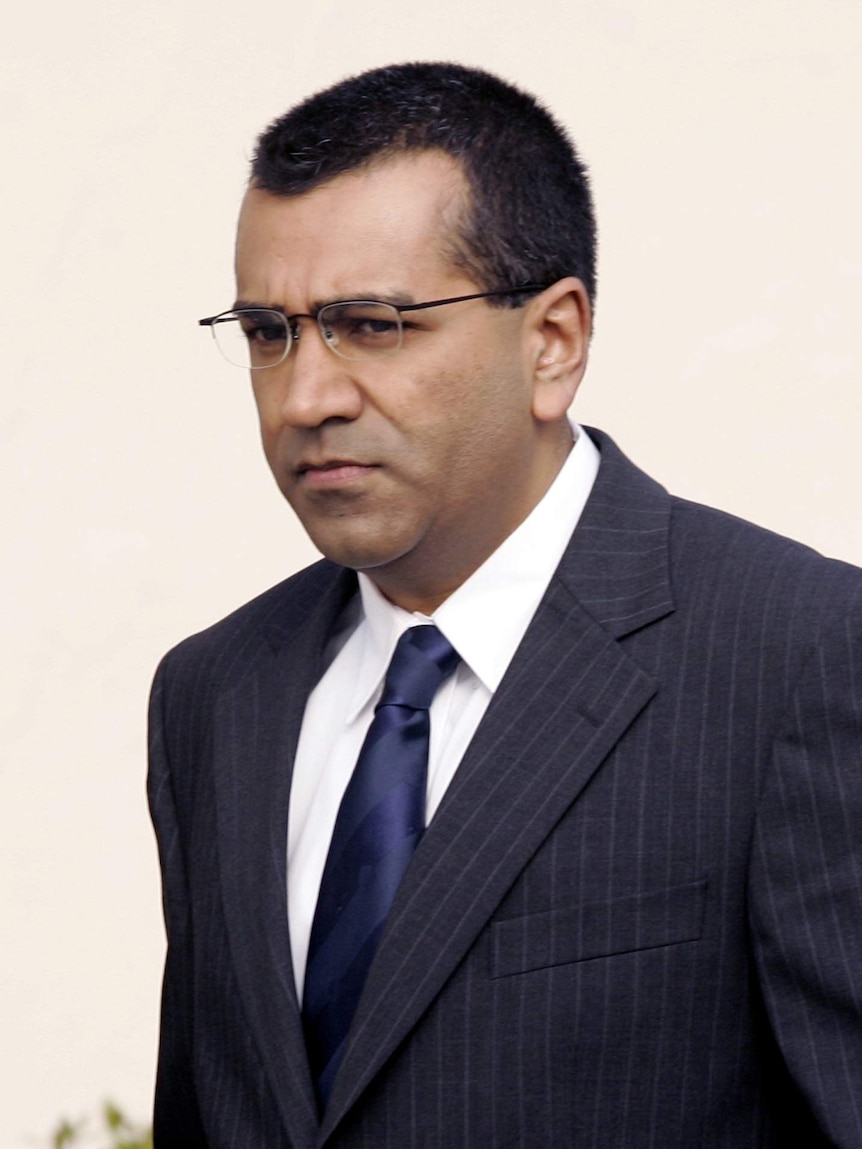 You view a portrait image of Martin Bashir in a dark suit, navy tie walking in front of a beige background.