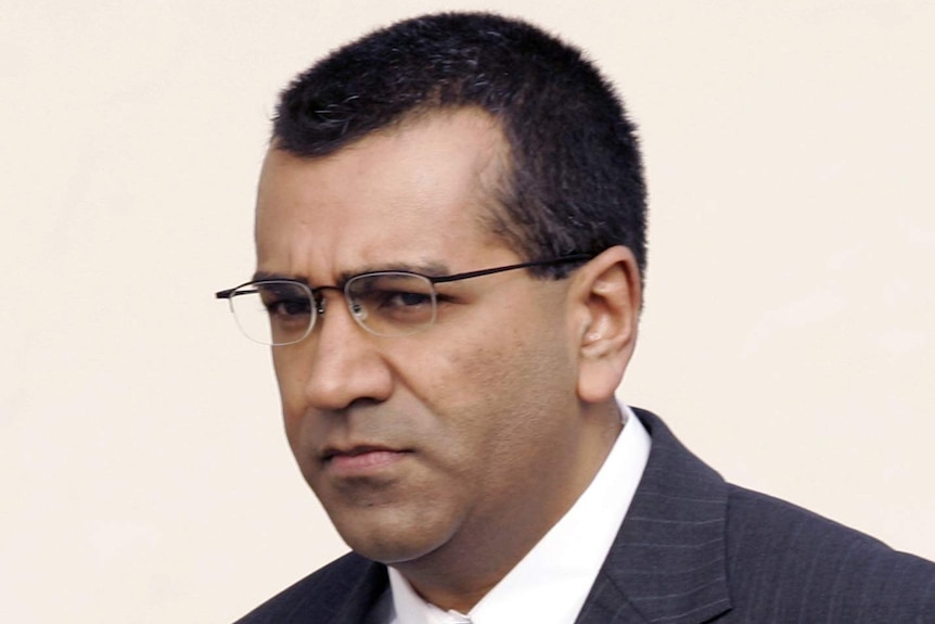 You view a portrait image of Martin Bashir in a dark suit, navy tie walking in front of a beige background.