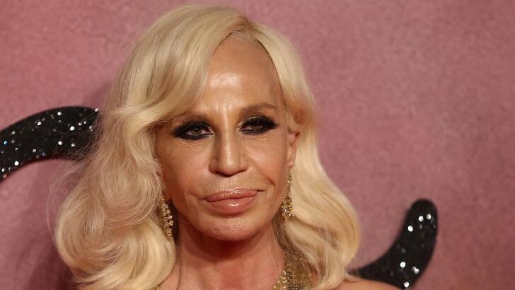Donatella Versace smiles with her mouth closed. She stands against a pink backdrop and wears a glomesh top.