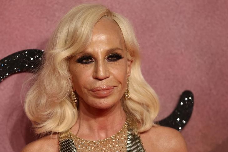 Donatella Versace smiles with her mouth closed. She stands against a pink backdrop and wears a glomesh top.