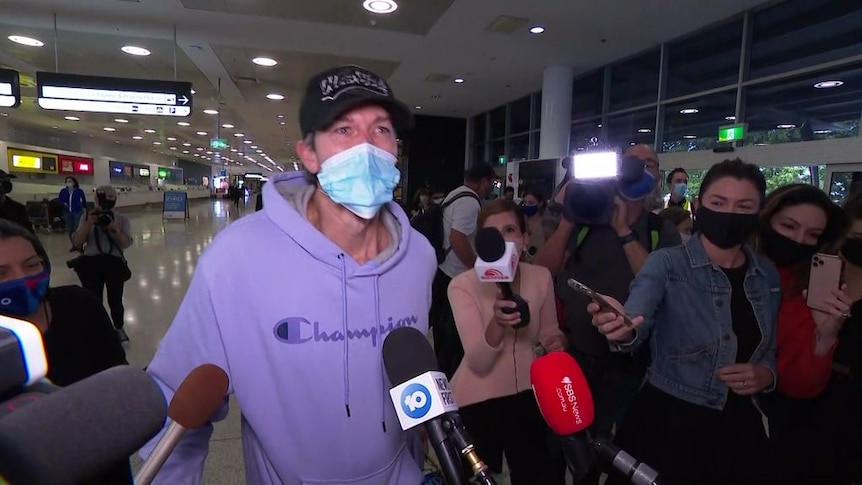 A man wearing a purple sweater and a face mask is being interviewed by news teams at Sydney Airport.