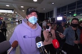 A man wearing a purple jumper and a face mask is interviewed by news crews at Sydney Airport.